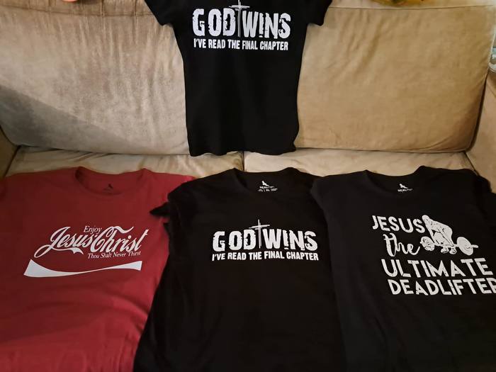 Four faith-themed t-shirts laid out, printed God Wins, Enjoy Jesus Christ and Jesus the Ultimate Deadlifter