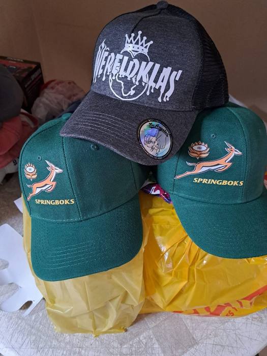 A grey trucker cap printed Wereldklas beside two green Springboks caps