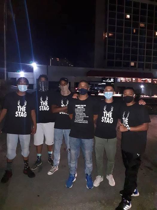 Six friends at a stag do in black The Stag t-shirts printed with antlers, one carrying the dare list